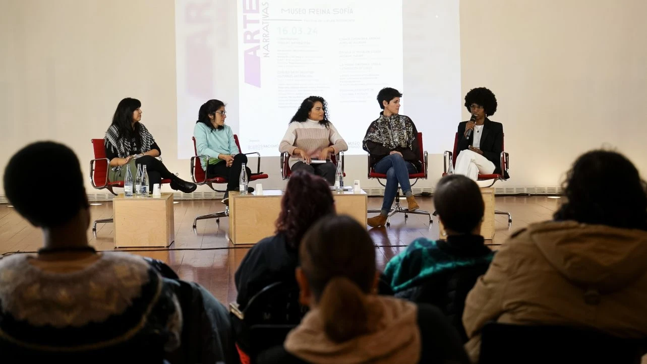 La imagen muestra a cinco mujeres de frente conversando en una sala bien iluminada y con público.