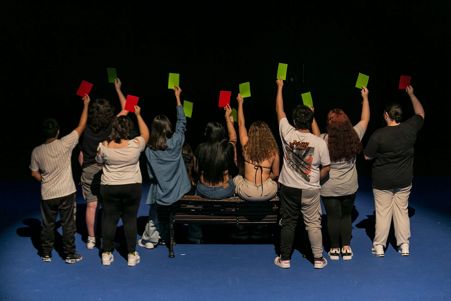 Diez personas sentadas en fila sobre un banco, vistas desde atrás, levantan tarjetas de colores en el aire —rojas y verdes— en un entorno con fondo oscuro y suelo azul