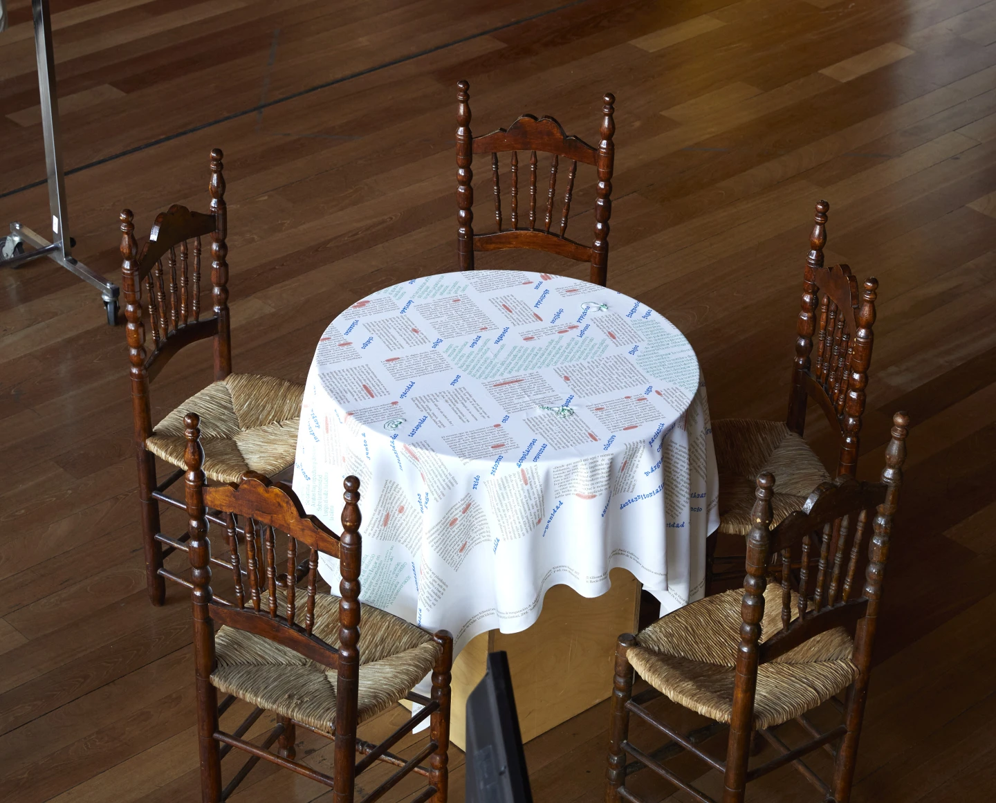 La imagen muestra una mesa redonda cubierta con un mantel blanco decorado con patrones coloridos y cuatro sillas de madera alrededor. El suelo es de madera y tiene un acabado brillante.