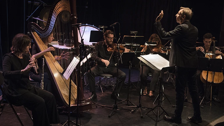 Varios músicos tocando instrumentos de cuerda, como el arpa, el violín y el violonchelo, frente a un director de orquesta