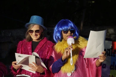 Aparecen dos mujeres en la imagen disfrazadas, ambas con gafas de sol, la primera lleva un sombrero azul y una chaqueta rosa brillante y sonríe mientras lee unos folios que lleva en la smanos. La segunda lleva una peluca azul brillante, una boa amarilla alrededor del cuello y lee una hoja de papel con una escobilla del baño como micrófono.