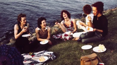 Fotografía en color que muestra a cinco personas en plano general, en un picnic al aire libre, sentadas en una pradera al lado del agua. Algunas de ellas son travestis. Están comiendo y riendo en un ambiente relajado y cercano. La imagen refleja una escena de la contracultura norteamericana del momento.