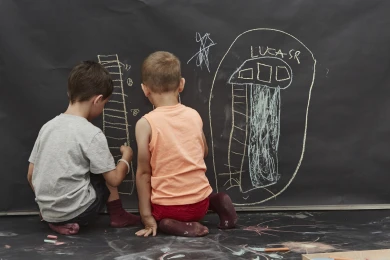 Dos niños sentados en sus rodillas pintan con tizas en una pared de pizarra
